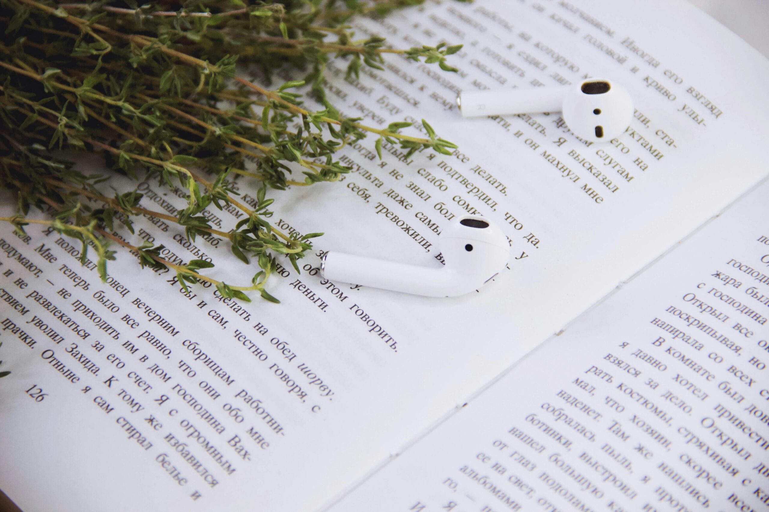 An open book with faint writing, white earbuds resting on the page, and green thyme leaves on the edge.